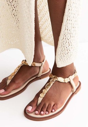 Stopy w beżowych płaskich sandałach z dekoracjami ze złotych monet i kremowej, szydełkowanej sukience.