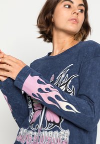 T-shirt de manga longa azul-marinho com padrão de chamas nas mangas, apresentando um design gráfico de uma borboleta e texto decorativo na parte da frente.