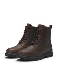 Par de botas de cuero marrón oscuro con cordones, suelas de goma negra y cremalleras laterales, inclinadas ligeramente hacia la derecha sobre un fondo blanco.