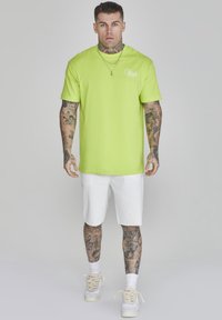 T-shirt in cotone verde neon con maniche corte, collo rotondo e dettaglio del logo. Abbinato a pantaloni corti in denim bianchi e sneakers bianchi.
