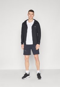 Melna uzvalka hoodie ar rāvējslēdzēju virs baltā T-kreklu, apvienots ar tumši ziliem šortiem ar rāvējslēdzējiem un melniem sporta apaviem.