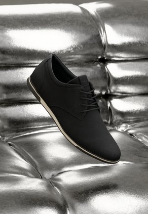Zwarte stoffen schoen met een gladde afwerking, vetersluiting en witte zool, zwevend boven een reflecterend metalen oppervlak.