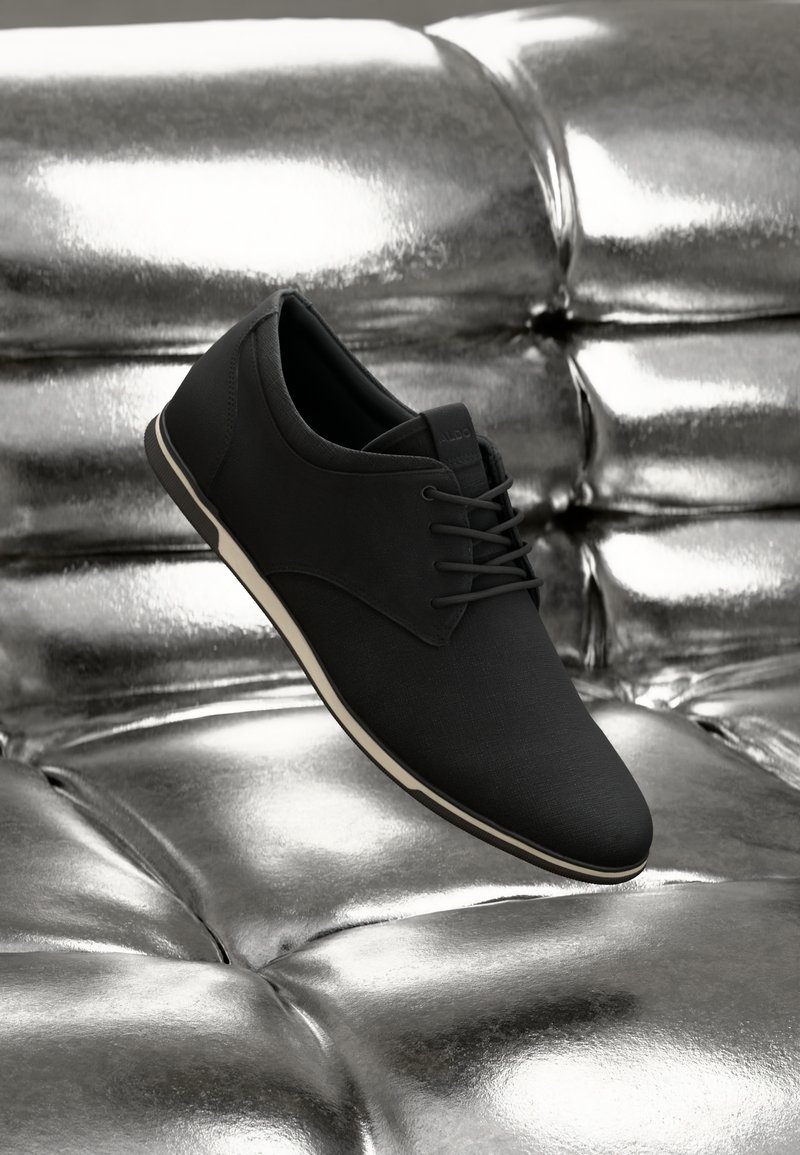 Zapato de tela negra con un acabado suave, diseño de cordones y suela blanca, flotando sobre una superficie metálica reflectante.