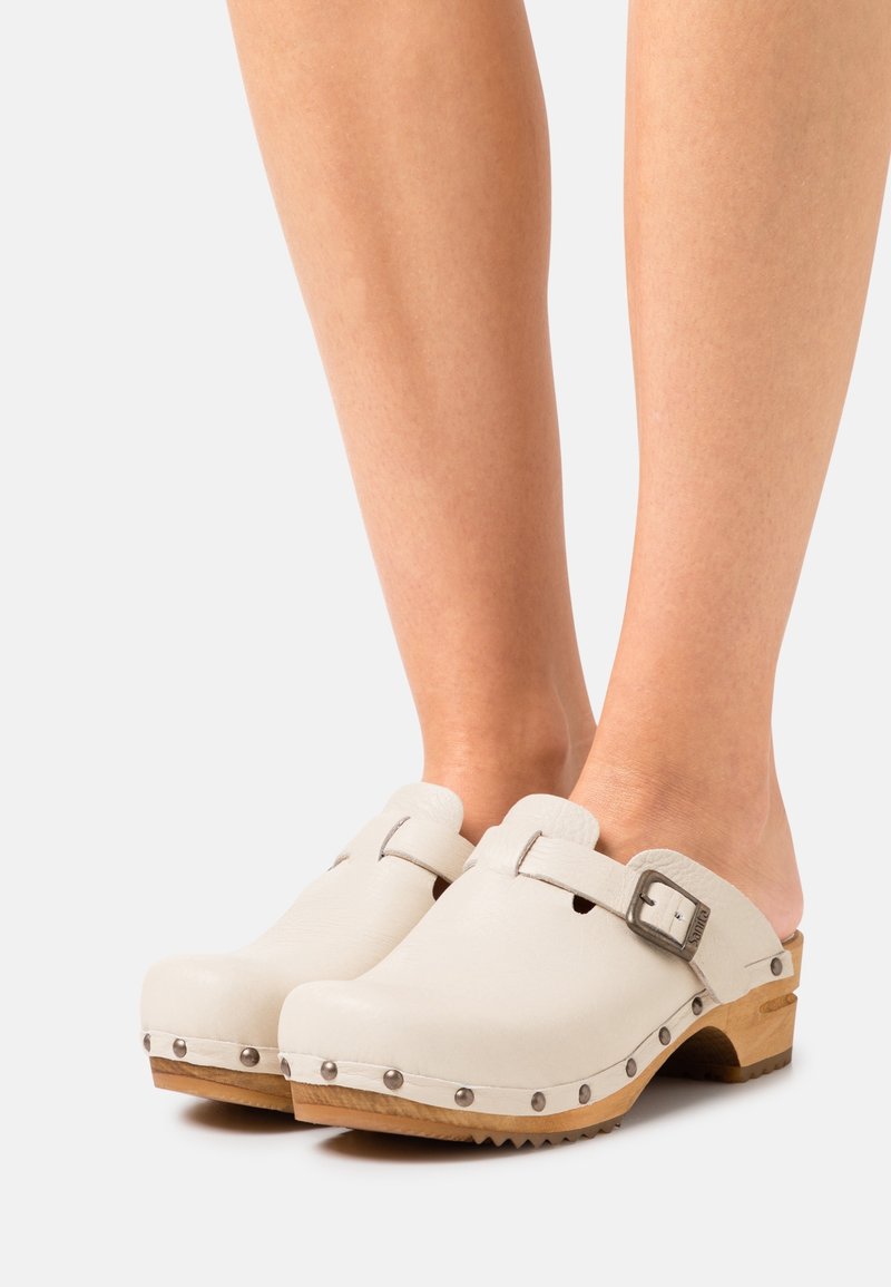 Jambes portant des sabots en cuir blanc avec semelles en bois et sangles à boucle argentée sur fond gris clair.