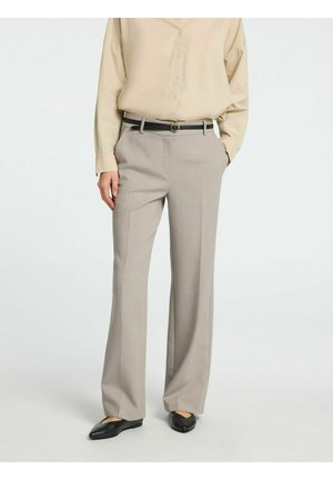 Personne portant une chemise beige à boutons rentrée dans un pantalon large gris clair avec une ceinture noire et des ballerines pointues noires, debout avec une main dans la poche.