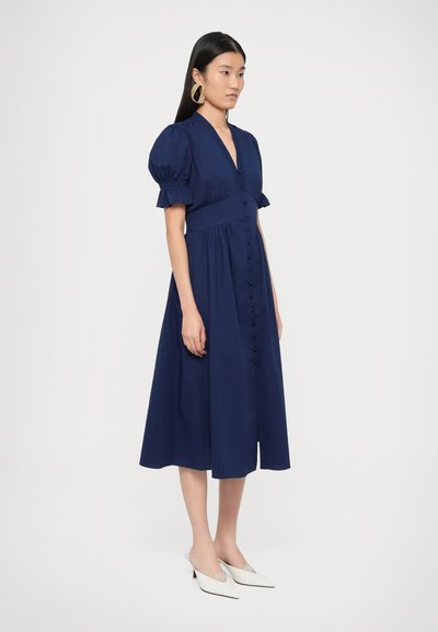 Φόρεμα σε ναυτικό μπλε με φουσκωτά μανίκια, V-λαιμόκοψη και εφαρμοστό κορσέ. Διαθέτει κουμπιά μπροστά και μήκος μέχρι τη μέση της γάμπας. Συνδυασμένο με λευκά παπούτσια με μυτερή μύτη.