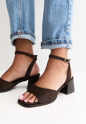 Bruine suède sandalen met blokhak en enkelbandjes, gedragen met omgeslagen lichtblauwe spijkerbroek op blote voeten.