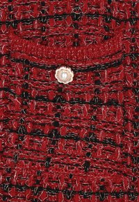 Piros kötött anyag fekete kiegészítőkkel és fehér szálakkal, texturált mintázattal. Egy díszes gyöngygomb van rávarrva a zsebre.