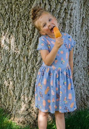 Junges Mädchen in einem blauen Kleid mit Eis-Muster leckt an einem orangefarbenen Eis am Stiel, während es draußen an einem großen Baumstamm steht.