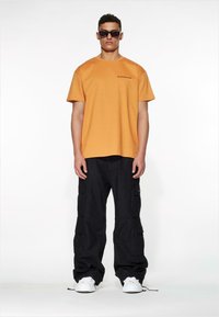 Oransje bomull T-skjorte med korte ermer, svarte cargo bukser med lommer, og hvite joggesko. Modell som bruker mørke solbriller.