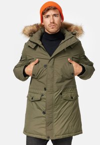 Parka verde oliva con capucha forrada de piel, cierres de botón y múltiples bolsillos. Presenta un exterior elegante y texturizado con un corte relajado.