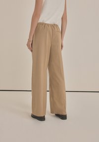 Pantaloni larghi color sabbia con righe verticali, vita elastica e una texture del tessuto liscia, abbinati a scarpe nere.