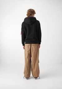 Felpa nera con cappuccio, pantaloni cargo beige a gamba larga con polsini elasticizzati, due tasche posteriori e un accentuazione rossa con zip sulla manica.
