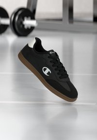 Cămașă sport neagră cu un material texturat, logo alb și talpă din cauciuc maro. Șireturile rotunde și gulerul căptușit îmbunătățesc confortul.