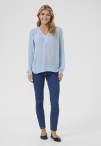 Blouse bleu clair à manches longues avec un décolleté en V et une texture subtile, associée à un jean en denim foncé ajusté et des chaussures plates noires.