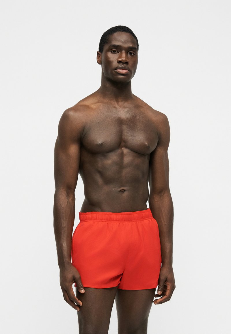 Muskeløs mand stående mod en ensfarvet baggrund iført lyse røde atletikshorts, kigger fremad med et neutralt udtryk.