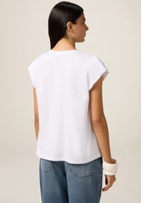 Vrouw met stijl Donker haar, dragend een losse witte top met korte mouwen en blauwe jeans, van achteren gezien, met stevige witte armbanden om haar pols.