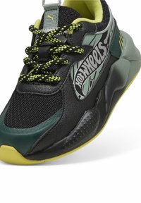 Chaussure de sport noire et verte en matières en maille et cuir, avec des accents jaunes vifs, une semelle texturée et un graphique "Hot Wheels" sur le côté.