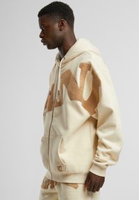 Beige zip-up hette med en lun brun "N" logo, kengurulomme og ribbestrikkede mansjetter. Laget av mykt materiale, med en avslappet passform.