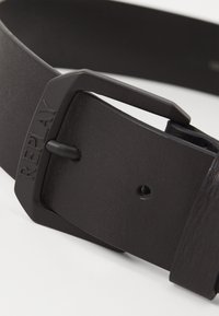 Sort læderbælte med en mat sort metalspænde med "REPLAY" præget. Teksturér overflade og flere justeringshuller synlige.
