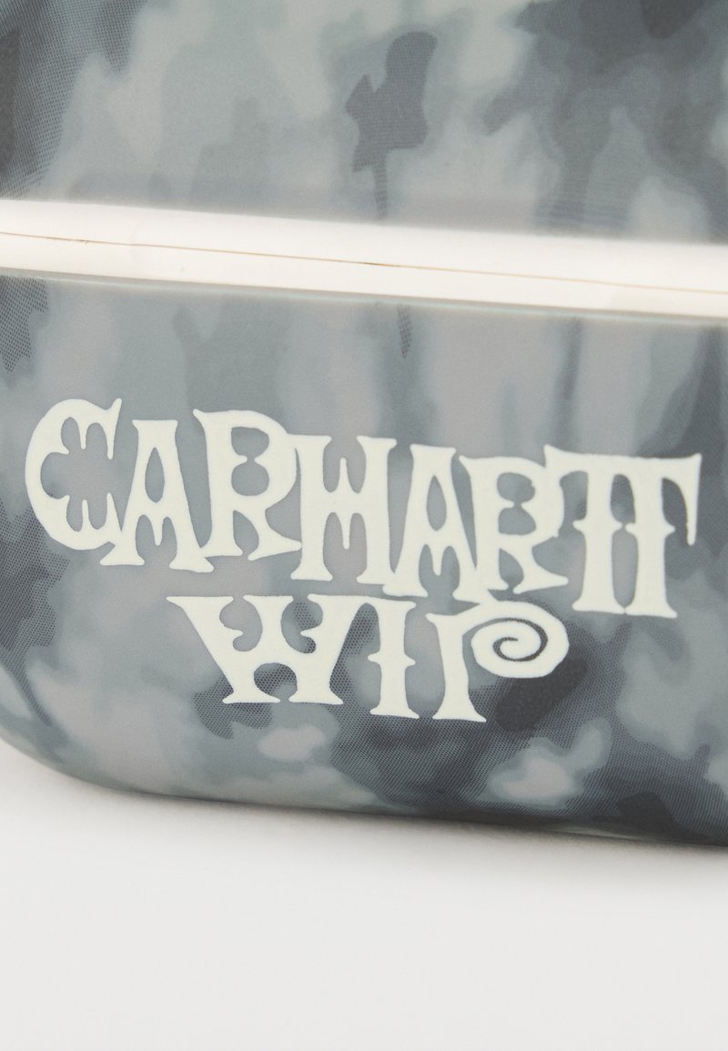 Gros plan sur un tissu camouflage gris et blanc avec une tirette blanche et le texte stylisé "Carhartt WIP" imprimé sous la fermeture éclair.