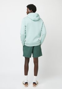 Felpa verde menta con tasca a marsupio, abbinata a pantaloni shorts verdi scuri. Il modello indossa calze bianche e scarpe da ginnastica con accenti marroni.