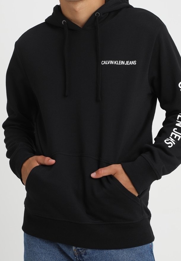 Sweat à capuche noir Calvin Klein Jeans avec poche avant et logo sur la poitrine et la manche gauche, porté avec un jean bleu, mains dans les poches.