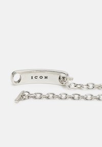 Pulseira de corrente em prata com uma etiqueta curva gravada com a inscrição "ICON." A corrente possui elos entrelaçados e um fecho tipo toggle.