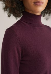 Burgunderfarbener Rollkragenpullover aus weichem, strukturiertem Stoff mit einer taillierten Passform, der einen hohen Kragen und Schulternahtdetails aufweist.