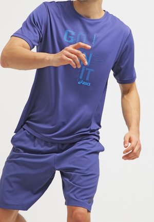 Homme portant un t-shirt et un short de sport Asics violet avec le texte "Go On It", posant comme s’il courait ou était en mouvement sur un fond uni.