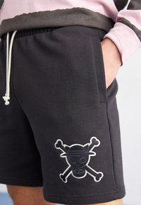 Shorts negros de tejido suave, con un logo bordado de calavera y huesos cruzados en blanco en la pierna. Cintura elástica con cordón.