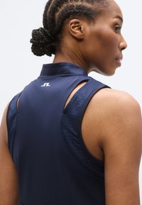 Tamsiai mėlynas sportinis be rankovių viršus su aukštu apykakle, dėmėtas orui pralaidžiais tinklais ant pečių ir nugaros, bei mažuoju baltu logotipu ties kaklu.