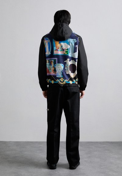 Schwarze Jacke mit gemustertem Rücken, der klassische Kunstmotive zeigt. Mit Kapuze, aus leichtem Stoff, kombiniert mit schwarzen Hosen und Schuhen.