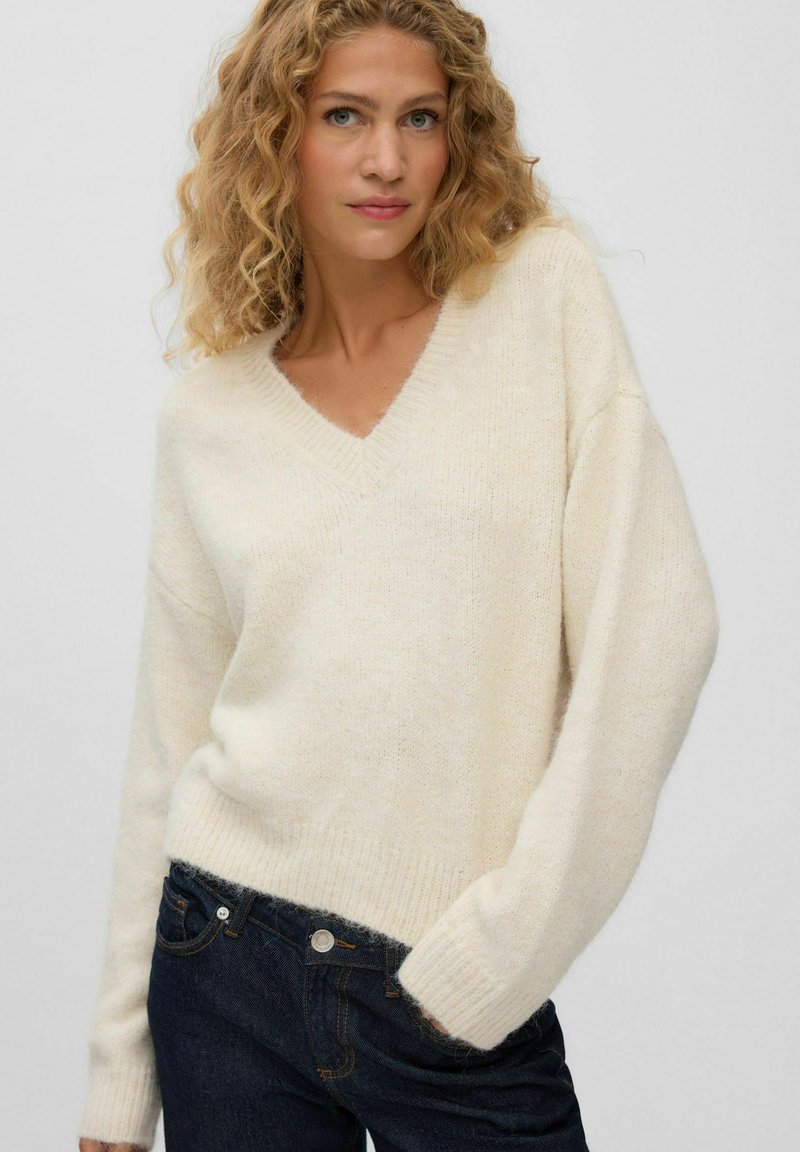 Femme aux cheveux blonds bouclés portant un pull crème à col en V et un jean bleu foncé, une main dans la poche, sur un fond clair uni.