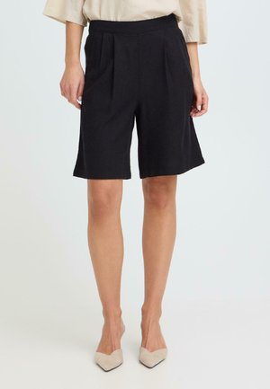 Kvinne iført svarte knelang plisserte shorts og beige spisse hæler, stående mot en ensfarget hvit bakgrunn.