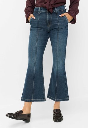 Kvinde iført mørkeblå udvidede jeans med forreste sømme, hænderne i lommerne, kombineret med mørkebrune loafers og en vinrød bluse.