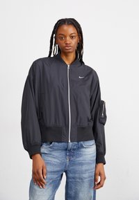 Ung kvinna med flätat hår som bär stora hoops örhängen, en svart zip-up Nike bomberjacka och ljusblå jeans mot en enfärgad bakgrund.