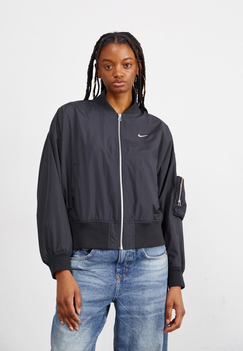 Ung kvinna med flätat hår som bär stora hoops örhängen, en svart zip-up Nike bomberjacka och ljusblå jeans mot en enfärgad bakgrund.