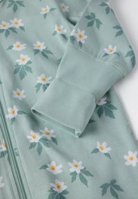 Lys grønne zip-up overall lavet af blødt stof, med et blomstermønster af hvide blomster med gule midter og grønne blade. Med manchetter ved ærmerne.