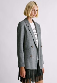 Donna bionda che indossa un blazer grigio doppiopetto, una t-shirt bianca con stampa e una gonna nera con frange, in piedi davanti a uno sfondo neutro.