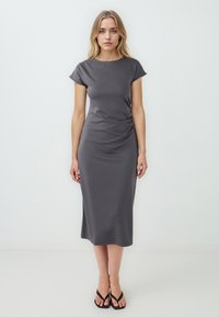 Robe grise jusqu'aux genoux avec des manches courtes et un col rond. Texture lisse et détail de fronces subtiles sur le côté. Portée avec des tongs noires.
