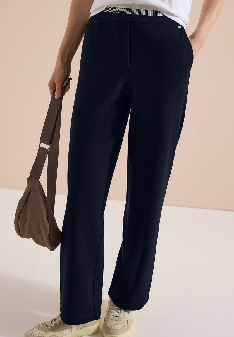 Persoon draagt marineblauwe wijde broek met gestreepte elastische tailleband, witte sneakers, draagt een bruine schoudertas met één hand in de zak.
