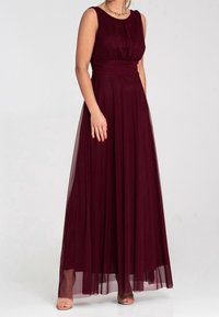 Femme portant une robe bordeaux sans manches longue jusqu'au sol, avec une taille cintrée et une jupe fluide, debout devant un fond clair uni.