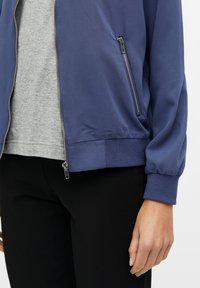 Marineblauwe zip bomberjack met ribbels aan de manchetten, zijzakken en een gladde textuur. Draagt over een lichtgrijs shirt, gecombineerd met zwarte broek.