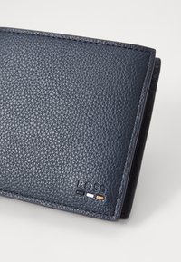 Carteira de couro azul-marinho com acabamento texturizado, apresentando um logótipo em relevo e três riscas de destaque em branco e laranja. Design dobrado.