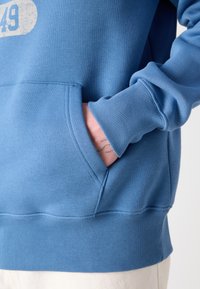Modra pulover z kapuco iz mehkega materiala z kenguru žepom, rebrastimi manšetami in robom. Ima zbledel tisk na prsih.