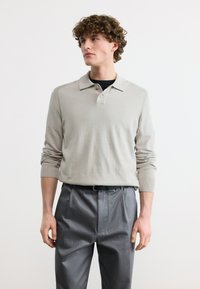 Νεαρός με σγουρά μαλλιά, ντυμένος με ανοιχτό μπεζ μακρυμάνικη πόλο μπλούζα και γκρι παντελόνι, στέκεται μπροστά σε απλό λευκό φόντο.