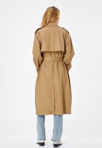 Mujer vestida con un abrigo trench beige con cinturón, jeans de mezclilla claros y zapatos de tacón blancos, de pie de espaldas sobre un fondo blanco.