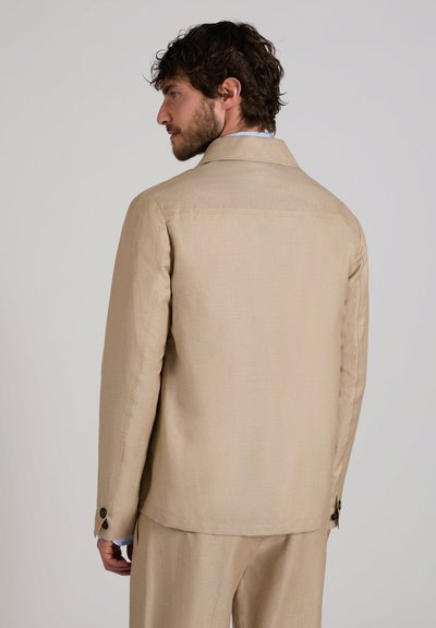 Hombre con pelo rizado que lleva una chaqueta de traje beige y pantalones a juego, visto desde atrás sobre un fondo neutro.