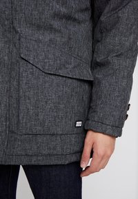 Chaqueta gris texturizada con un puño abotonado, costuras visibles y una etiqueta de logotipo en el área del bolsillo inferior. Primer plano de la manga y el bolsillo.